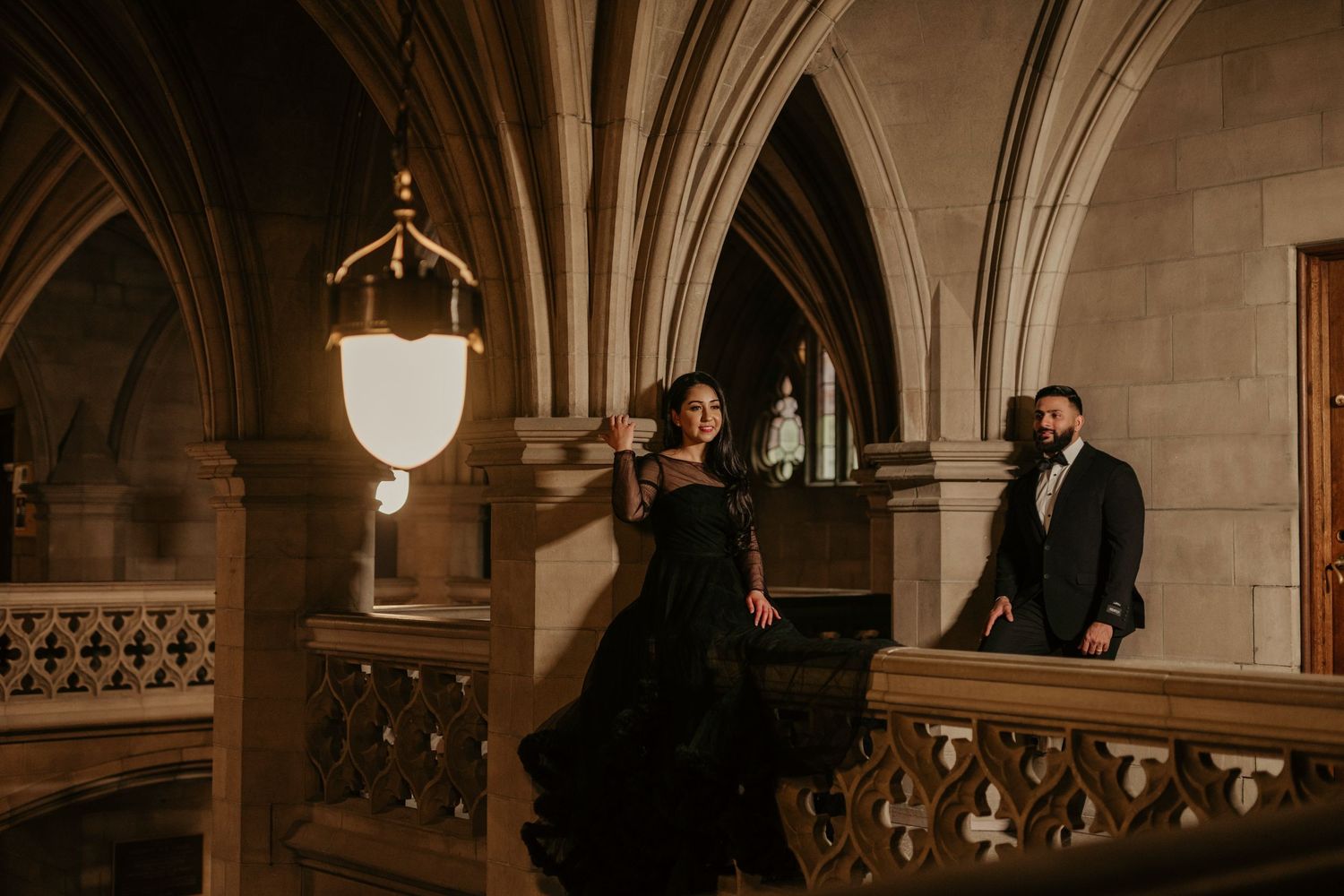 A couple in elegant black attire poses in a Gothic-style interior with arched ceilings and ornate architectural details.