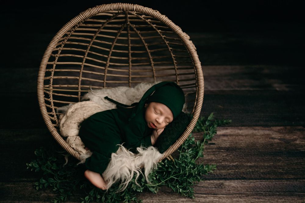 Small person wearing dark winter clothing rests peacefully in a woven basket chair surrounded by greenery.