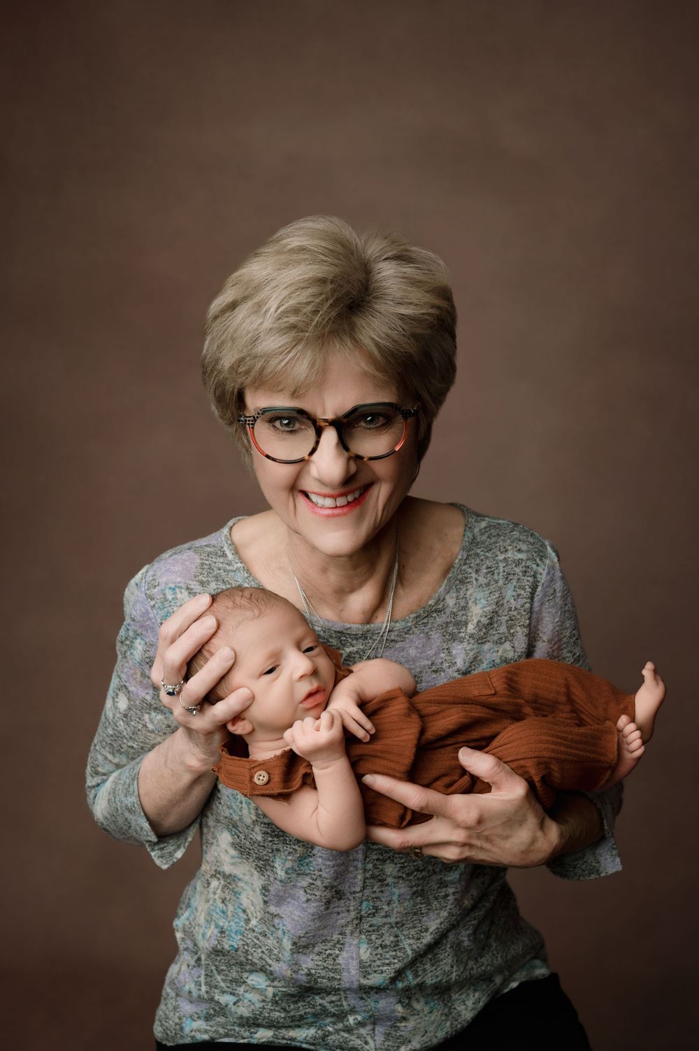 A woman in glasses and gray floral top holds a sleeping newborn baby wrapped in a brown outfit in a studio portrait.