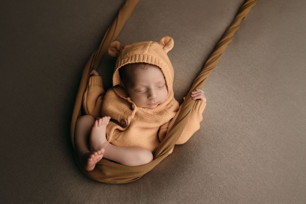 A sleeping newborn in a knit hat lies nestled in a beige fabric sling.