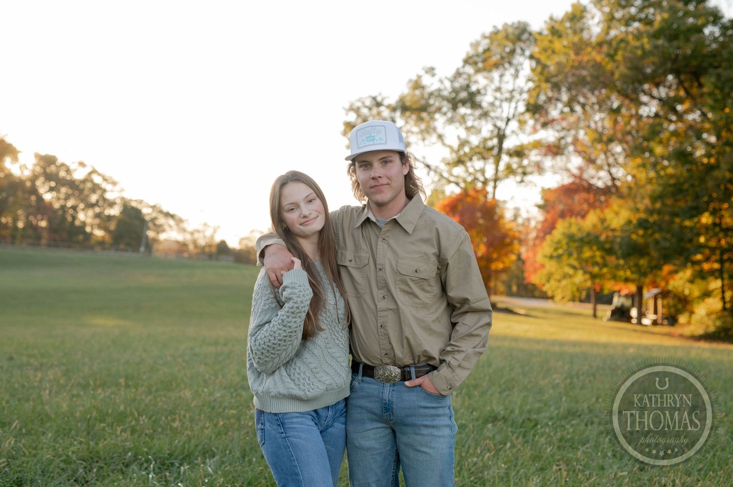 A couple stands close together in a sunny field with golden fall trees behind them.