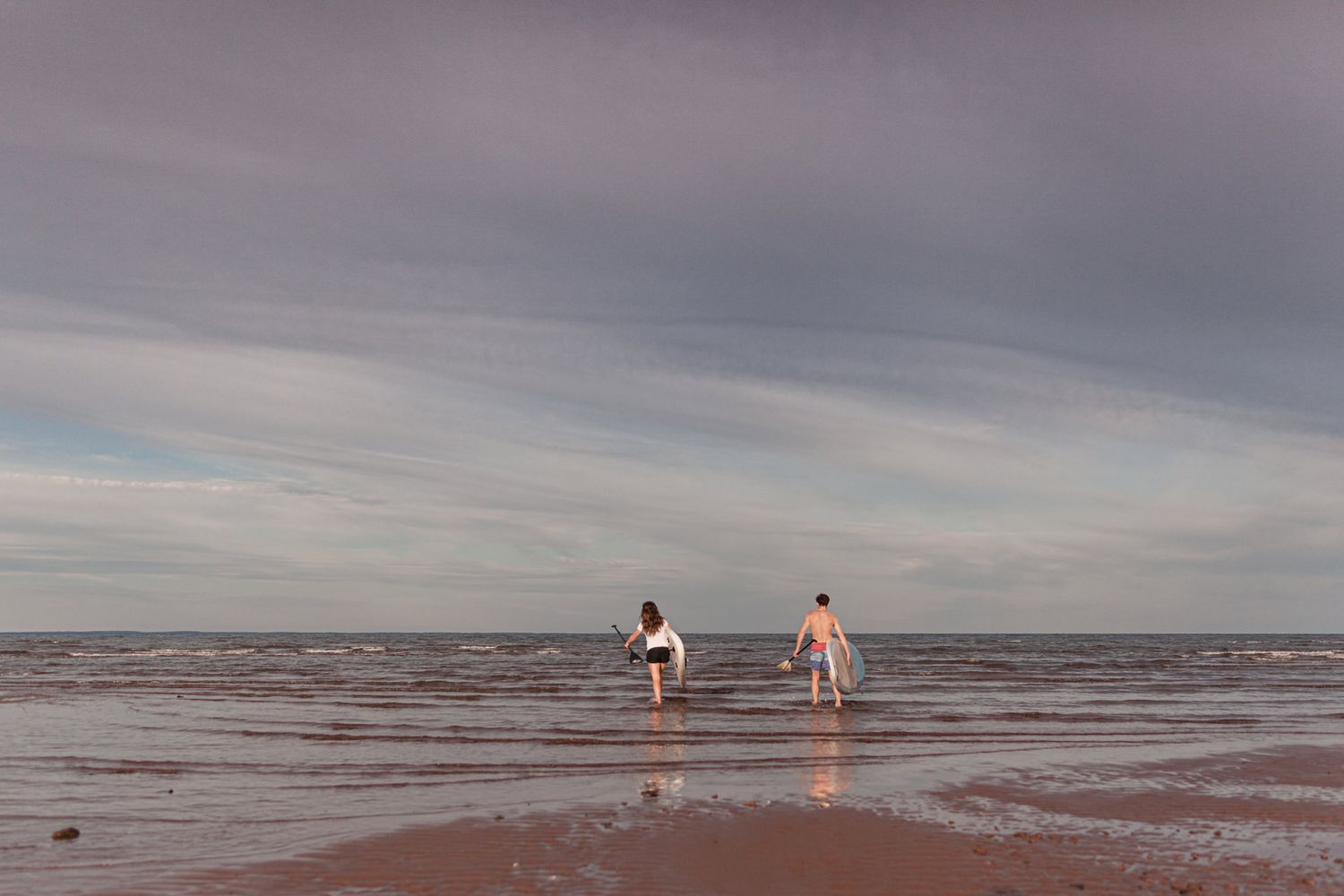 Couple carrying paddleboards on a beach during a cinematic couples photoshoot in Chelton, PEI Canada with Lumenali Photography