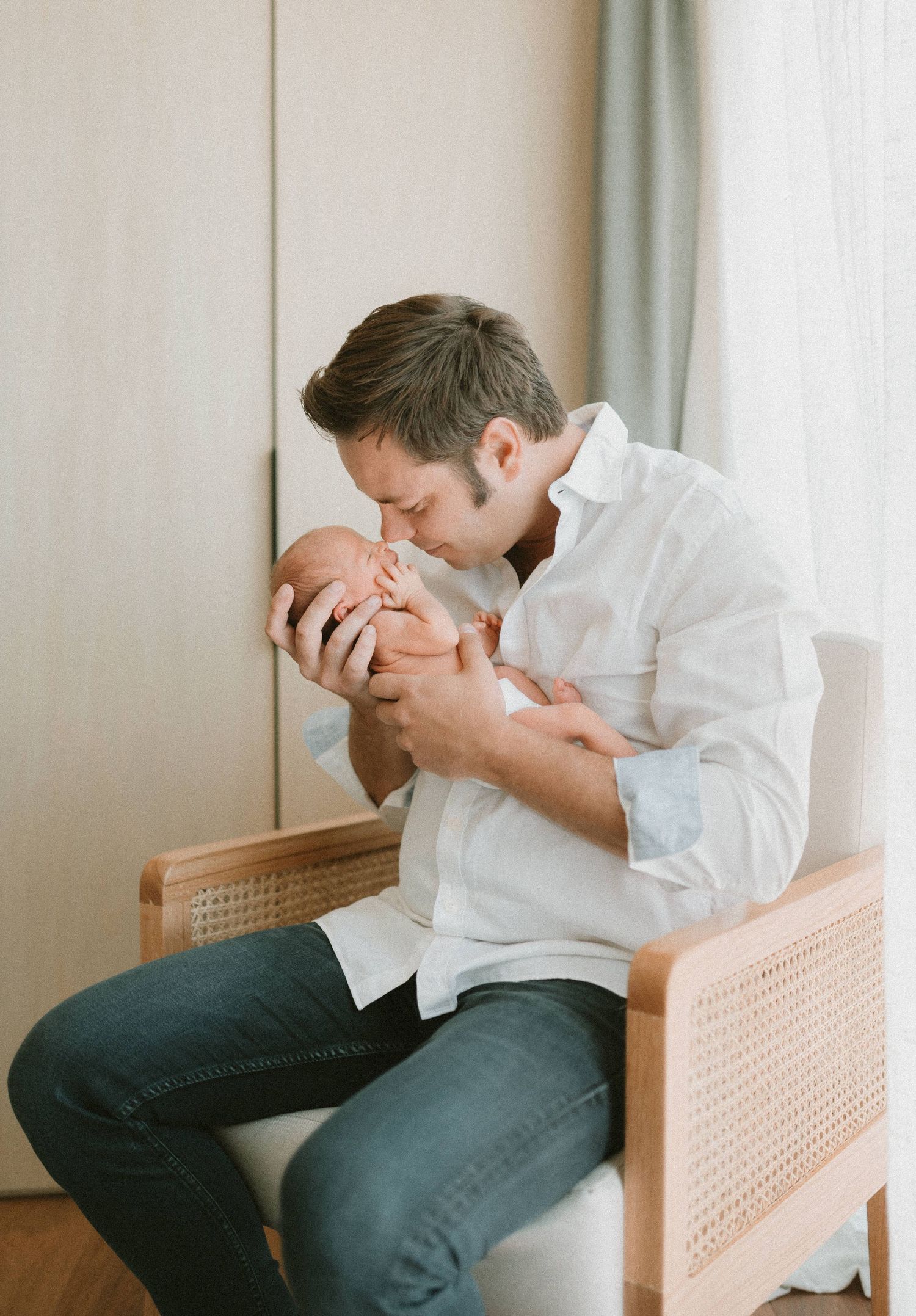 A parent in a white button-down shirt tenderly holds and kisses a newborn baby while sitting in a cane-backed chair.