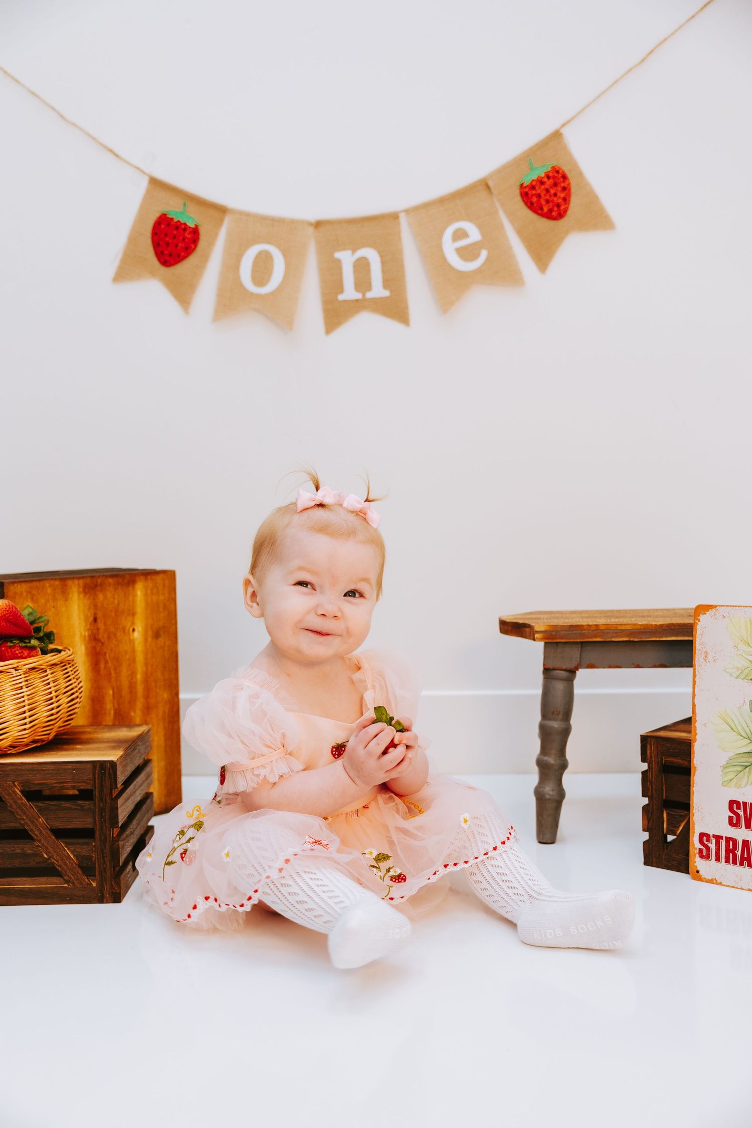 A first birthday celebration setup with a burlap banner and strawberry decorations against a white backdrop.