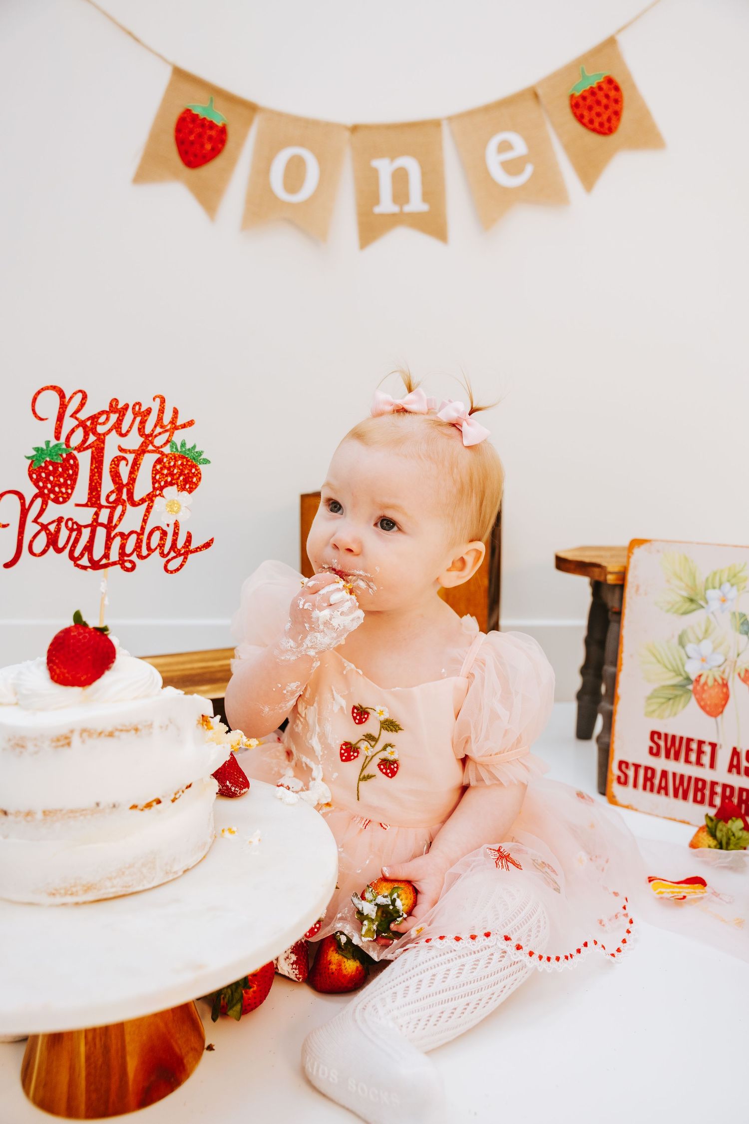 A first birthday party scene with a white cake and strawberry-themed decorations.