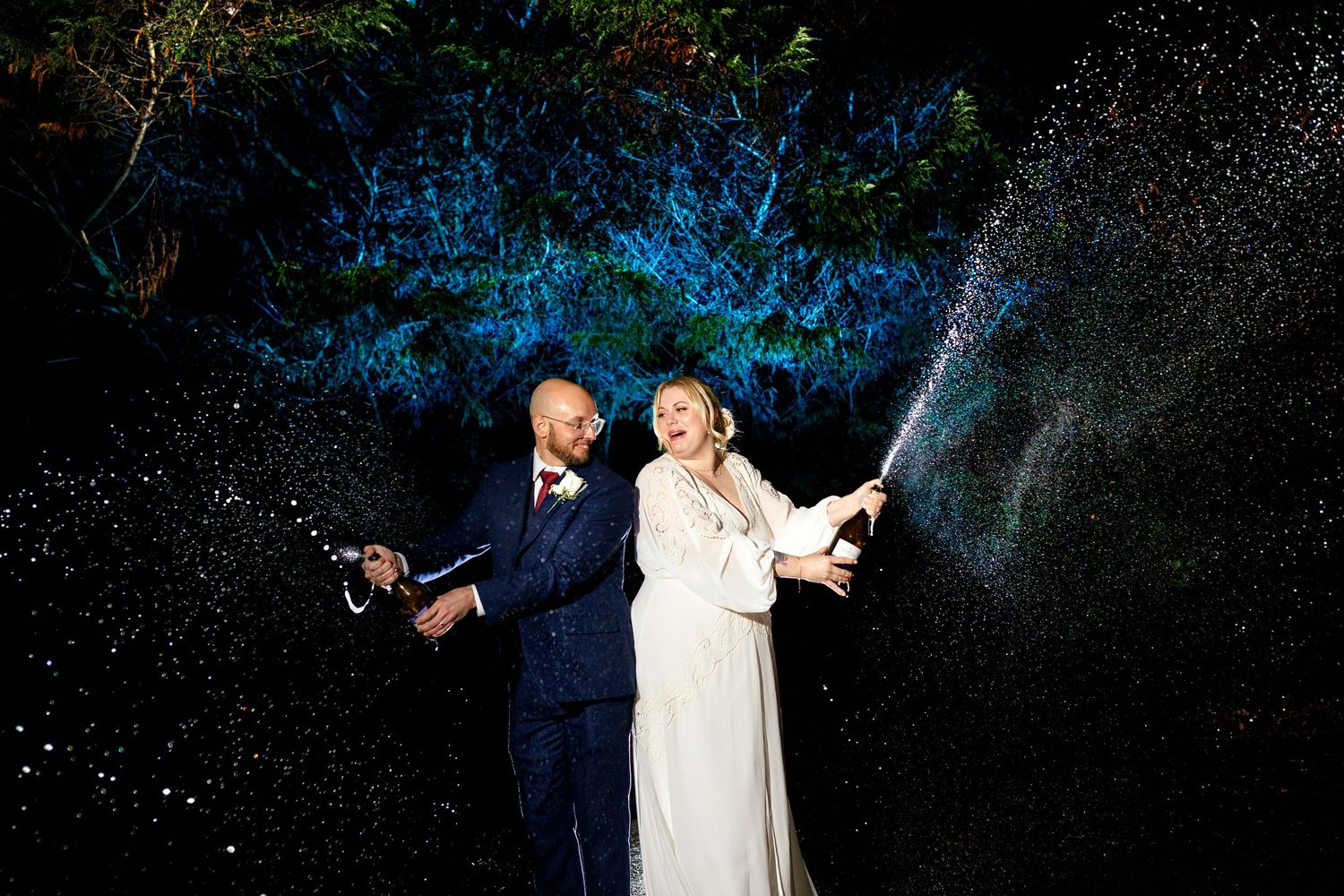 A couple in wedding attire sprays champagne at night against a dramatic blue-lit background creating magical sparkles.