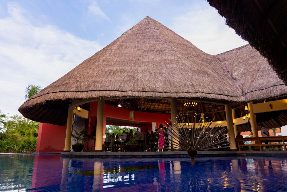 Tropical resort pool area with traditional thatched-roof palapa and red walls reflecting in blue water.