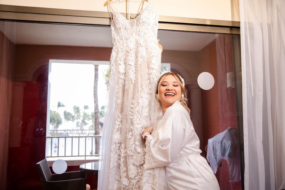 A white lace wedding dress hangs in a hotel room with morning light streaming through windows.