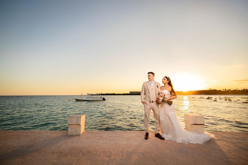 A couple in wedding attire stands on a pier overlooking the ocean at sunset as golden light illuminates the horizon.
