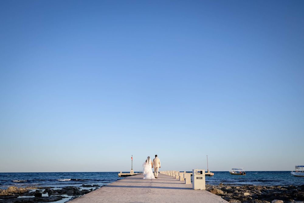 A couple walks on a long ocean pier surrounded by blue waters on a bright sunny day in Hawaii.