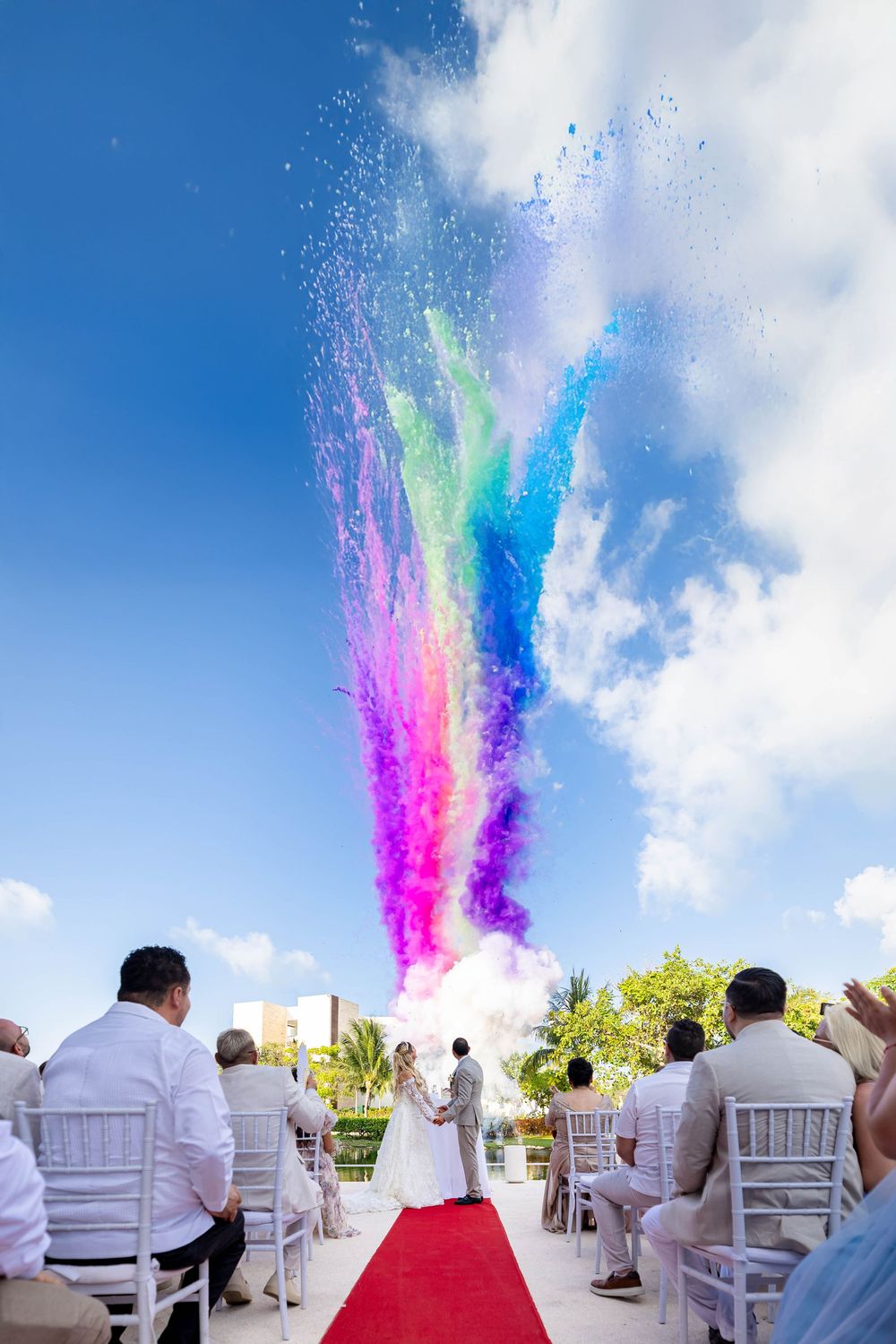 Colorful explosion of rainbow powder burst fills the sky during an outdoor wedding ceremony with red carpet and white chairs.