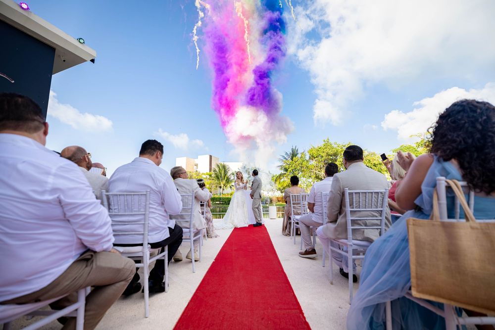 Pink smoke floats above guests seated in white chairs at an outdoor ceremony along a red carpet aisle.
