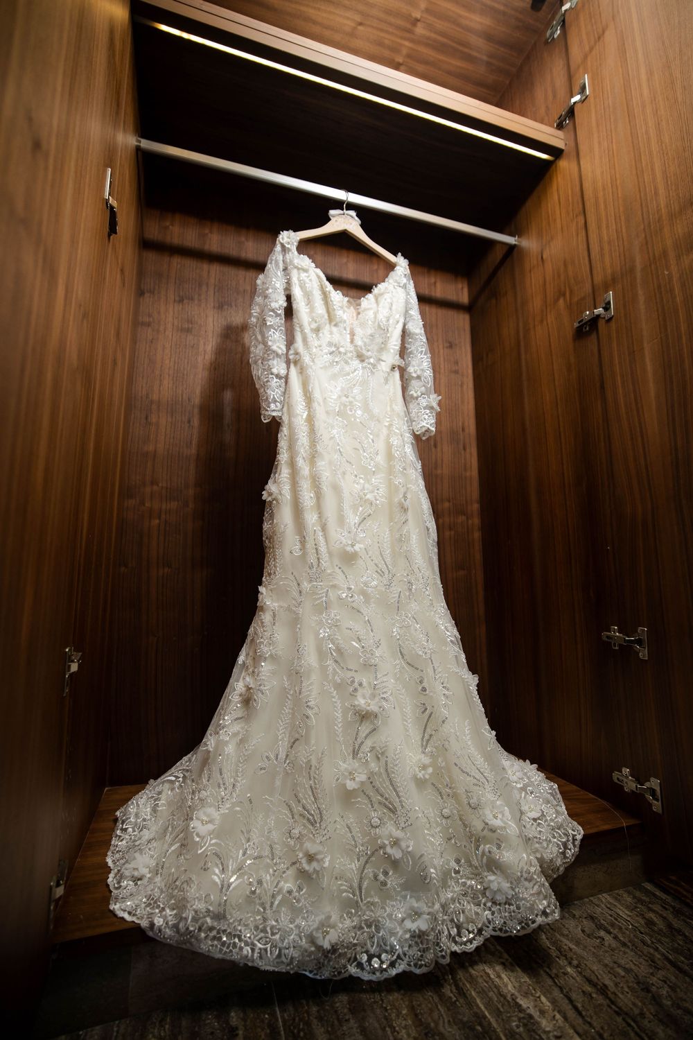 White lace wedding dress with intricate embroidery details hanging in wooden wardrobe closet.