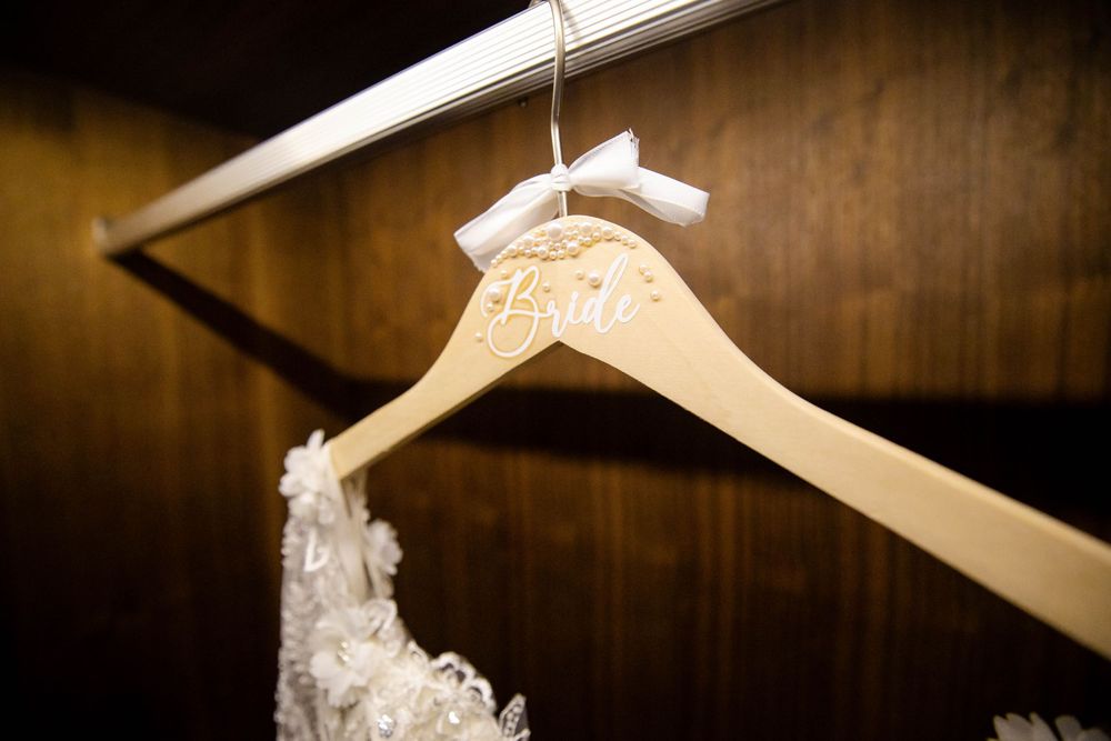 A white lace wedding dress detail hangs on a personalized wooden hanger against a dark wooden background.