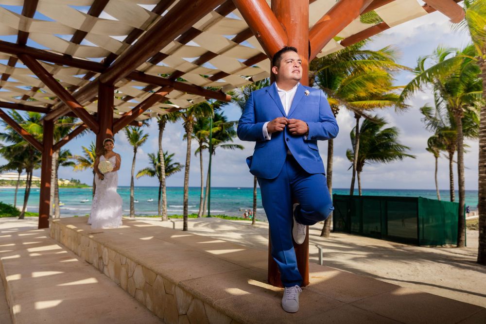A groom in a blue suit poses under a covered beachfront walkway with palm trees and ocean views.