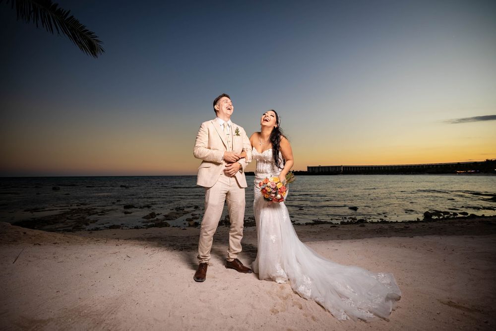 Newlyweds share joyful moment on beach at sunset with ocean and white sand in background.