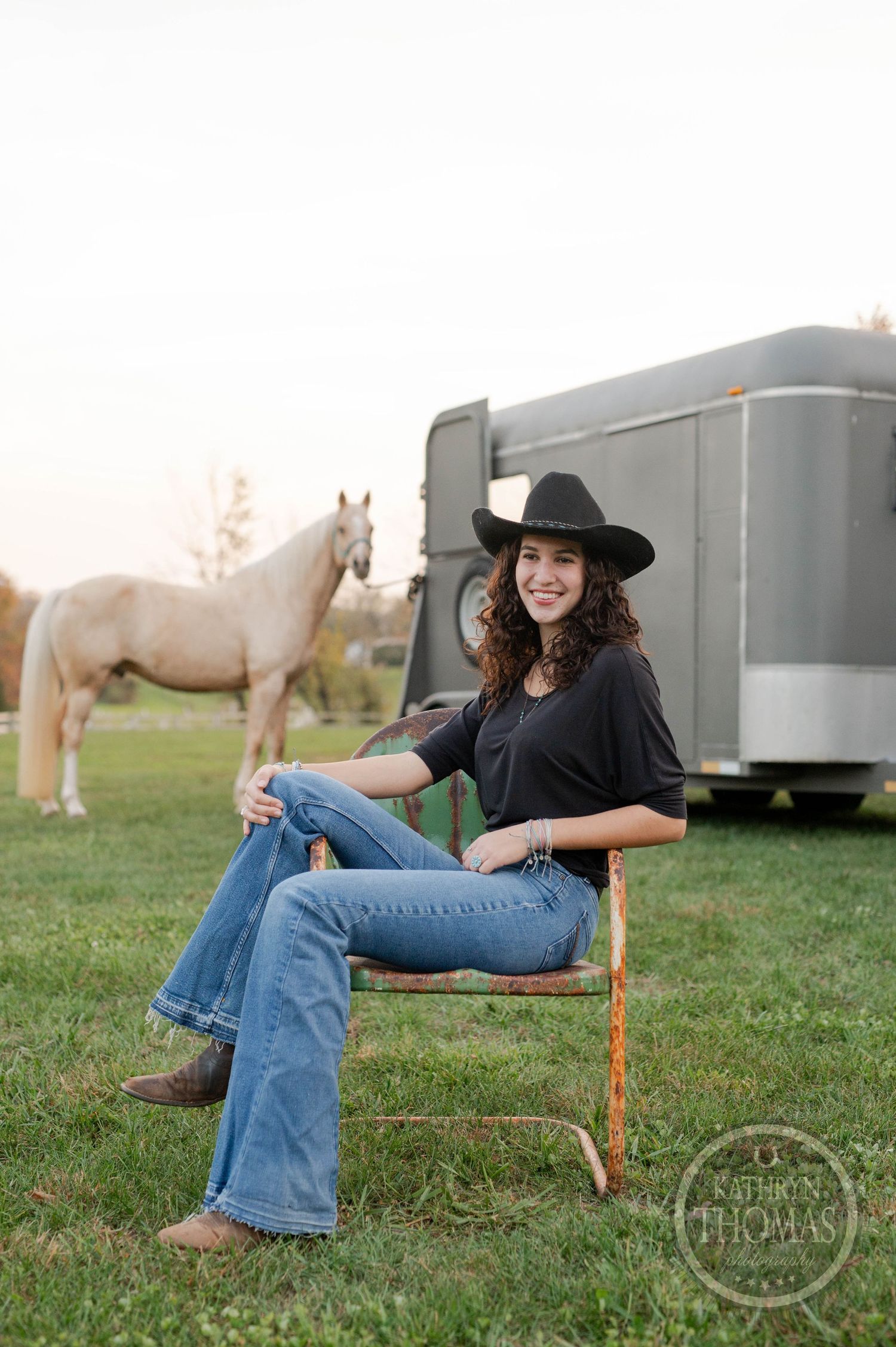 A relaxed western-styled portrait with a white horse and horse trailer in the background on a grassy field at sunset.