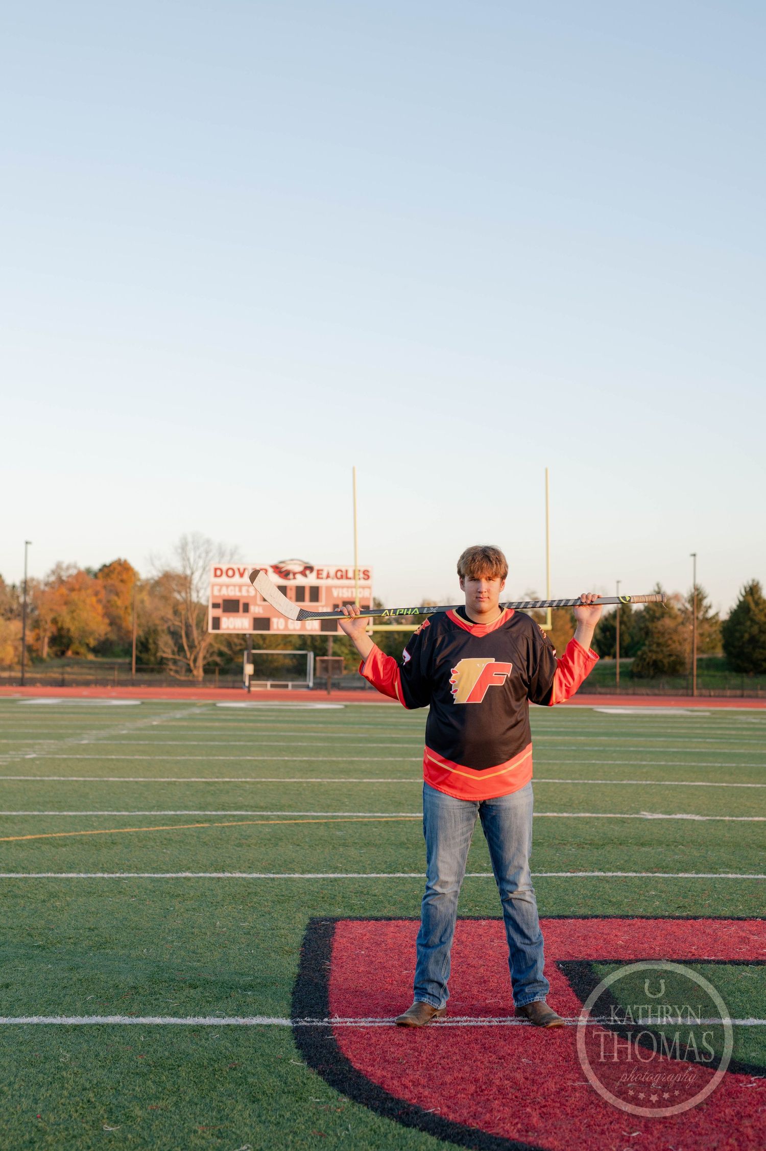 Player stands on football field holding hockey stick with stadium in background.