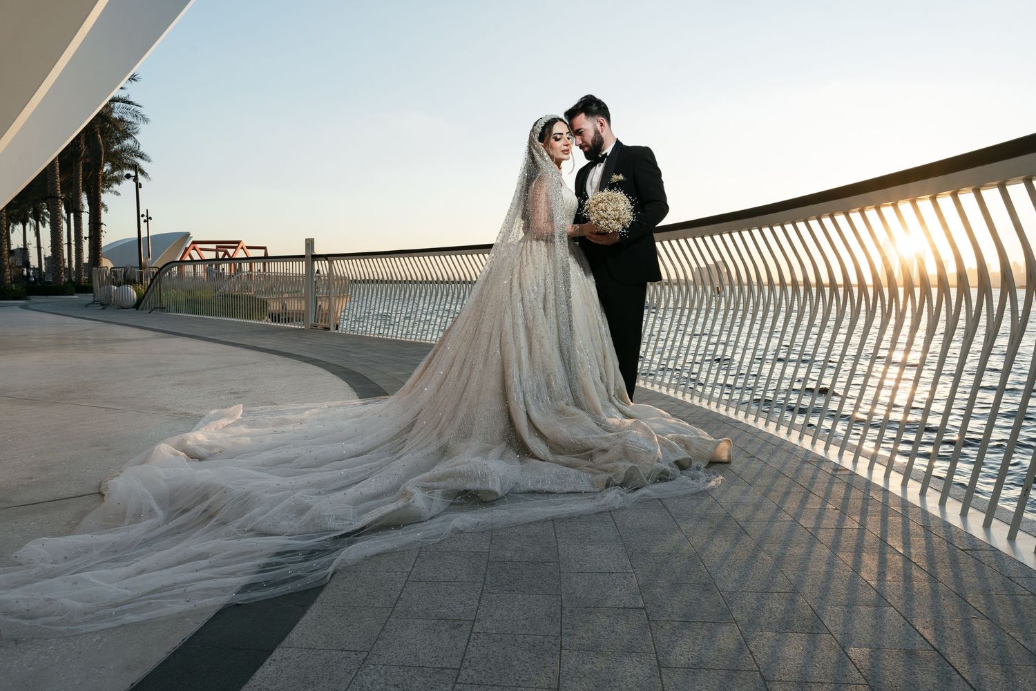 A couple in wedding attire pose on a waterfront promenade at sunset with a flowing cathedral veil trailing behind.