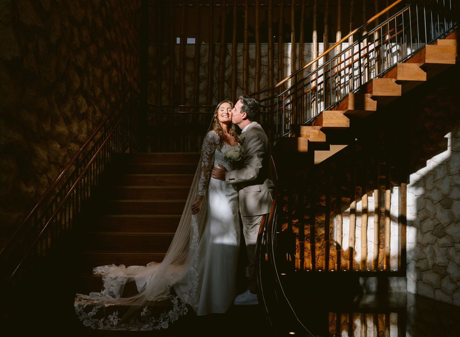 A couple in wedding attire pose on a waterfront promenade at sunset with a flowing cathedral veil trailing behind.