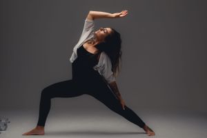 A dancer in black leggings and gray top performs an expressive contemporary dance pose against a dark studio backdrop.