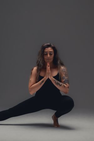 A person in dark athletic wear performs a deep yoga squat with hands in prayer position against a gray background.