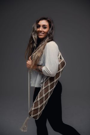 Cheerful person in a light colored top and black pants poses with a macrame yoga mat bag against a dark background.
