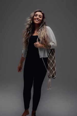 A person wearing a black jumpsuit and gray cardigan stands casually with a macrame bag against a dark background.