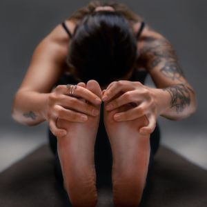 Person in black athletic wear doing a seated forward bend yoga position, focusing on the feet and legs against dark background.