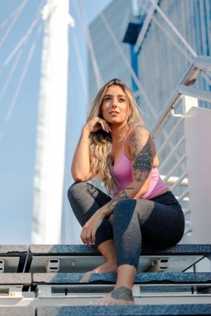 Athletic woman in pink sports bra and leggings sits on bleachers with modern architecture and blue sky in background.