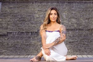 A person in a white dress sits on a stone patio with visible tattoos and long wavy hair.