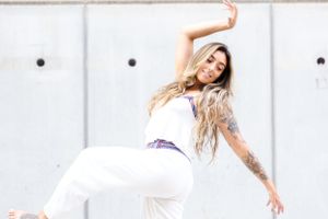 A dancer in white pants and tank top strikes an expressive pose against a white wall during a dance performance.