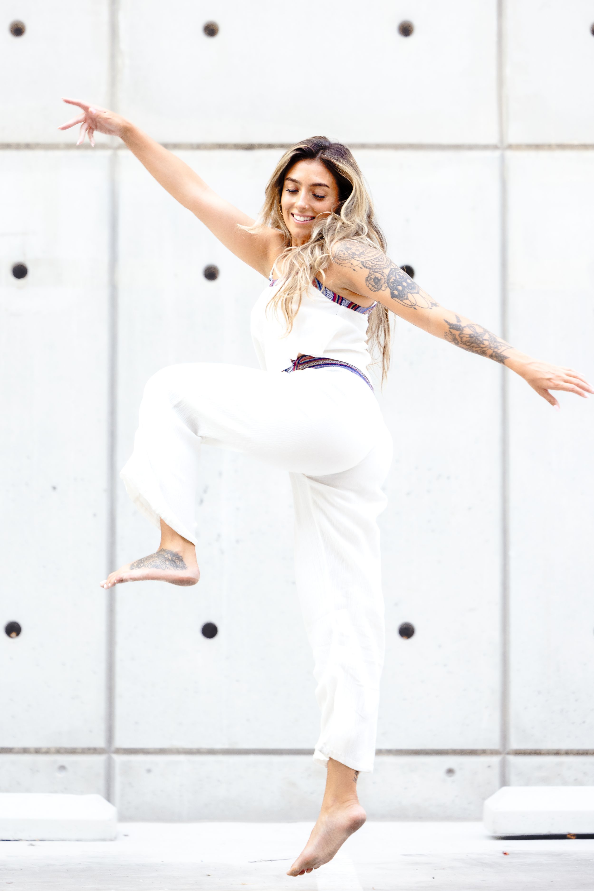 Woman in flowing white outfit dances gracefully against concrete wall background, arms outstretched in elegant pose.