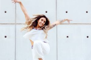 Dancer in flowing white outfit leaps joyfully against modern gray paneled wall.