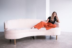 Stylish curved cream sofa with wooden legs and person lounging in bright orange pants on minimalist grey backdrop.