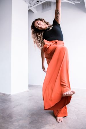 A dancer in a coral maxi skirt and black top strikes an elegant pose against a white wall while barefoot.
