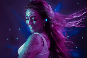 A dancer in glowing body paint performs under neon purple and blue lighting with flowing hair and ethereal effects.