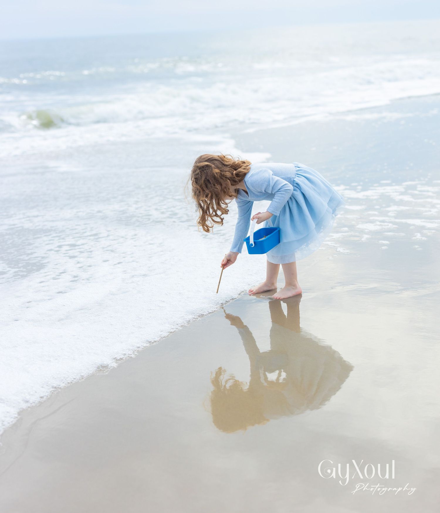 A child in a light blue dress draws in the wet sand at the ocean's edge while their reflection appears in the water.