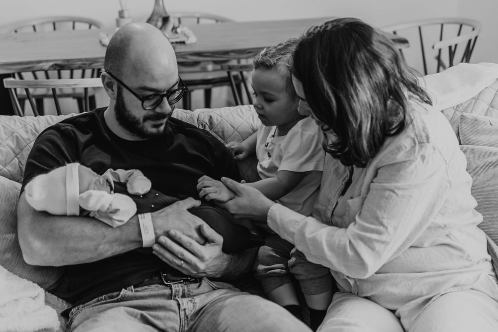 dad holding baby sister and mom showing to the oldest son how to touch the baby carefully, a day after she was born