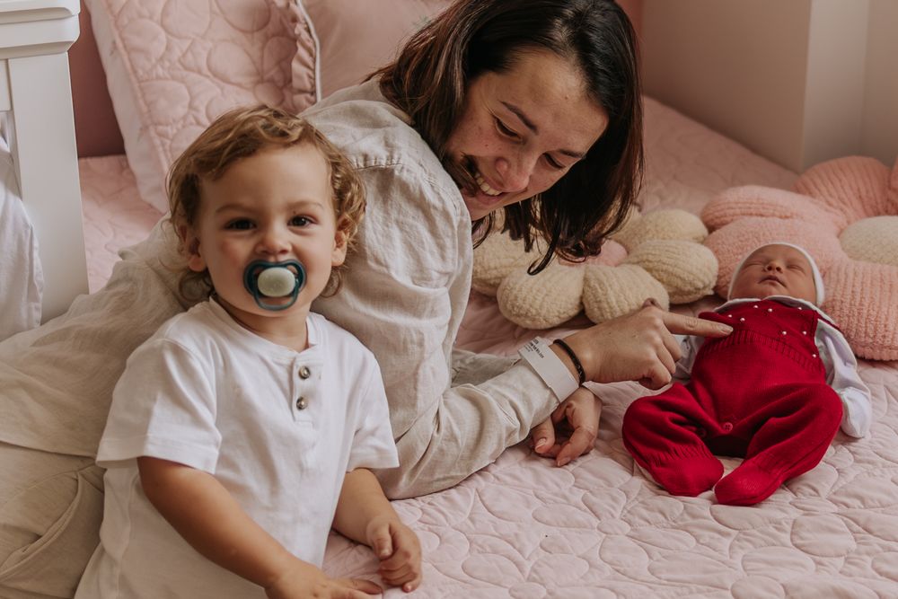 big brother looking proud to the camera while his mom shows him his baby sister in the bad