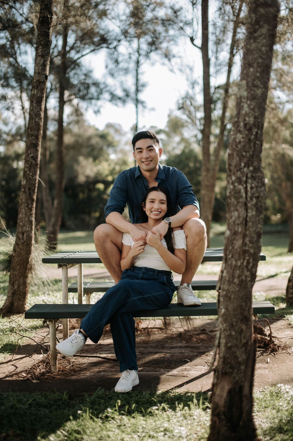 A couple poses together on a wooden picnic bench surrounded by tall trees in a sunlit forest.