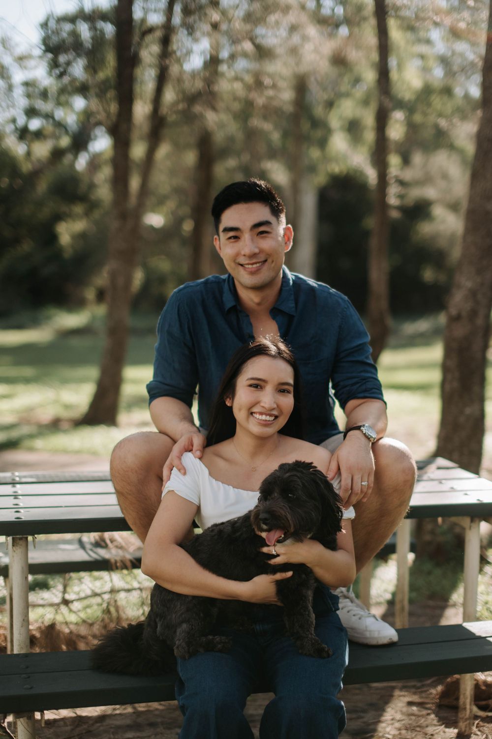 A couple sits together on a wooden bench in a leafy park with their small black dog.