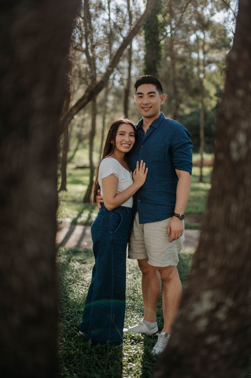 A couple stands close together in a wooded park setting wearing casual attire.