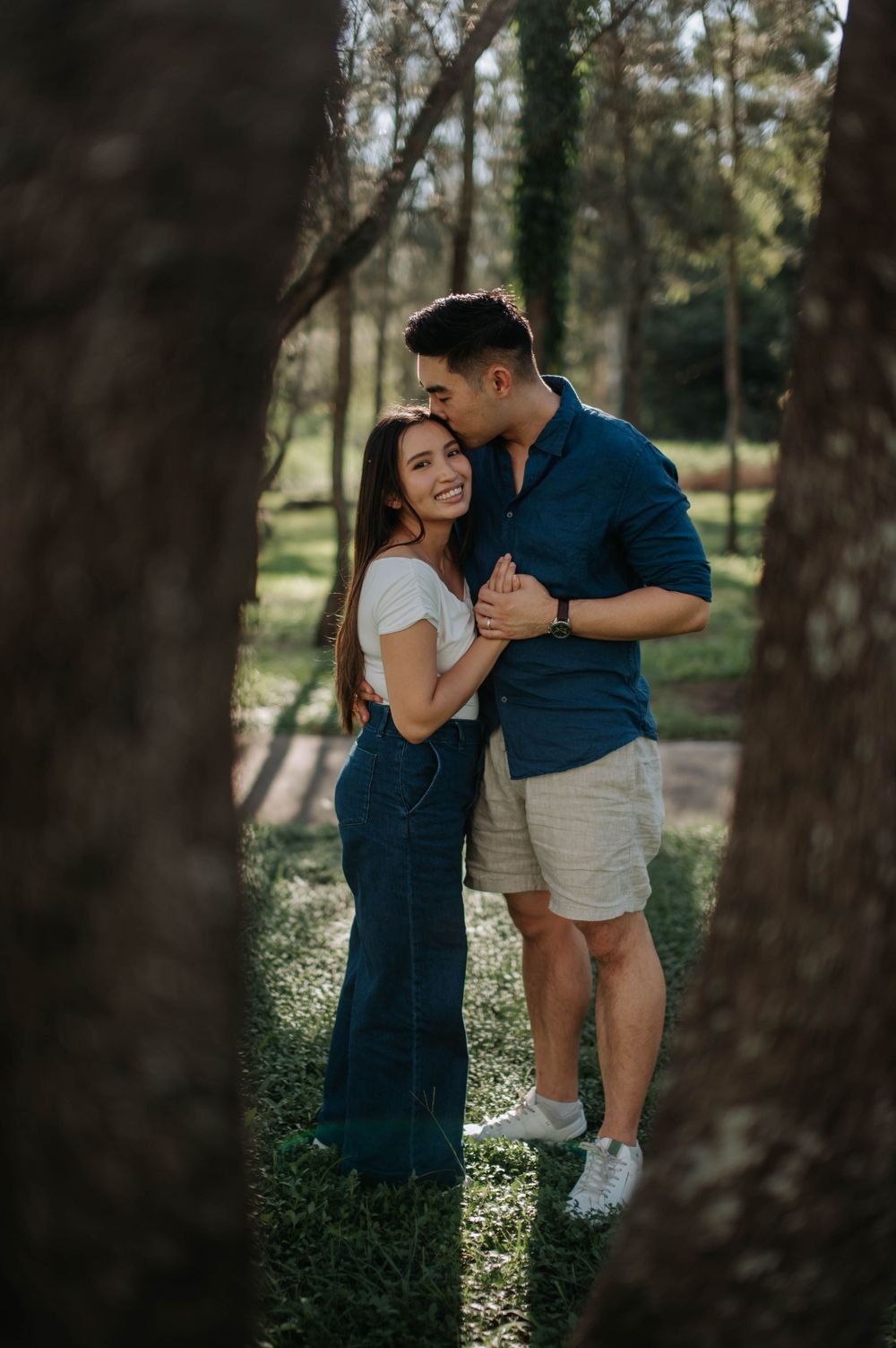 A romantic couple embraces while standing between trees in a sunlit forest setting.