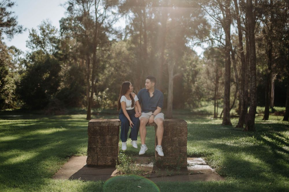 A couple sits together on a stone structure in a sunlit park with trees in the background.
