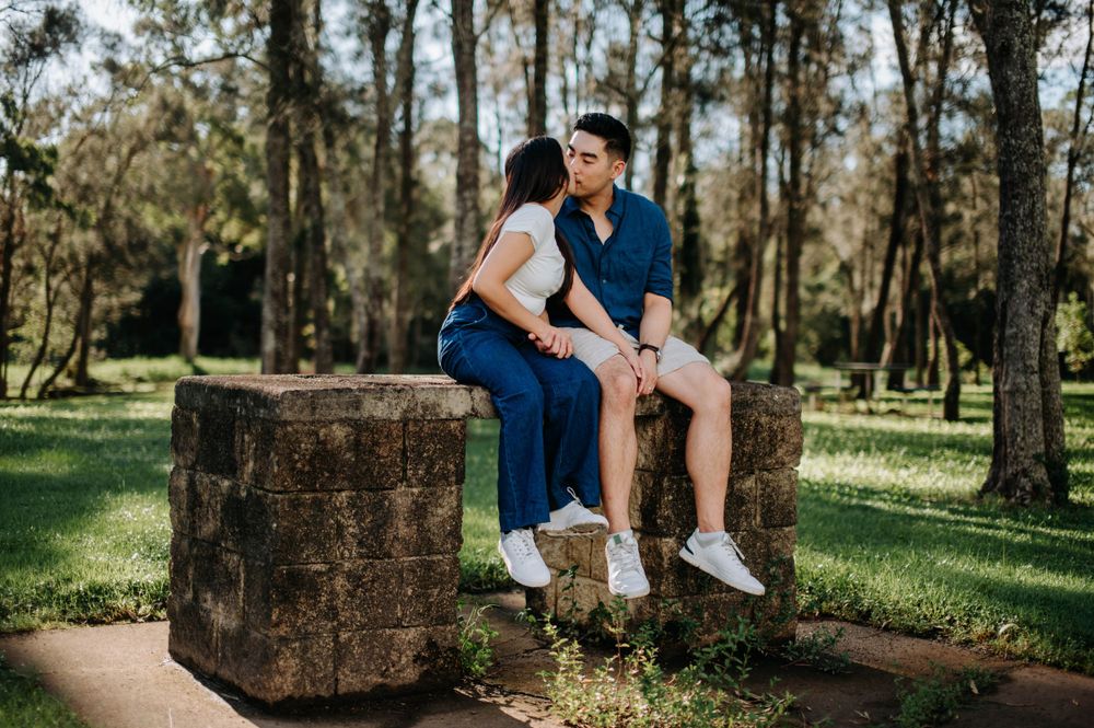 A couple shares a tender moment while sitting on a stone wall in a forest setting.