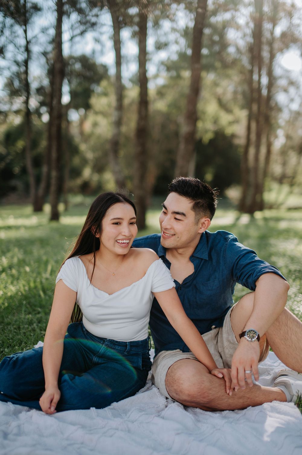 A couple relaxes together on a blanket in a sunny wooded park setting.
