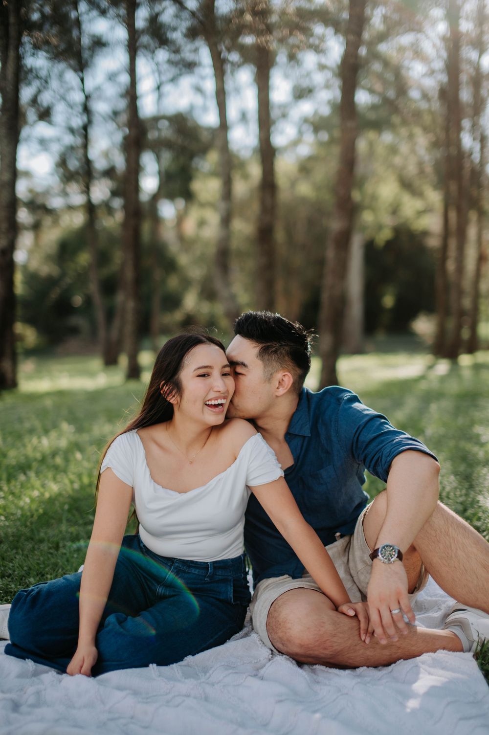 A romantic couple sharing a tender moment while sitting on a picnic blanket in a sunlit forest setting.