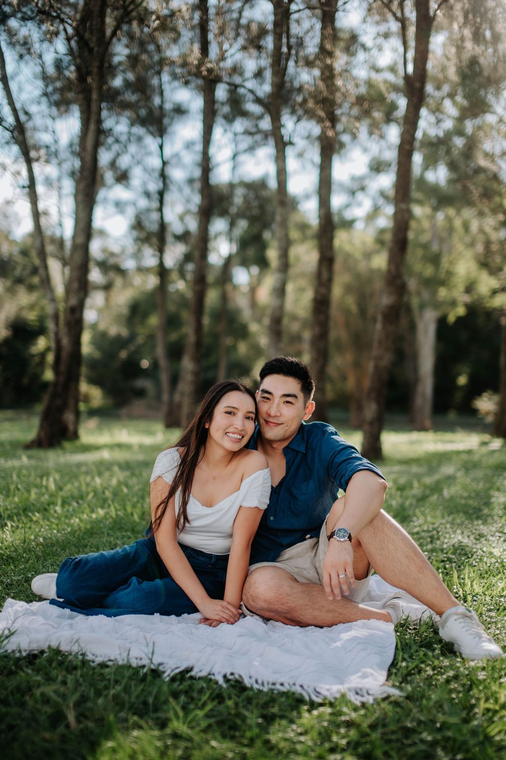 A couple relaxes together on a white blanket during an outdoor photoshoot in a wooded park.