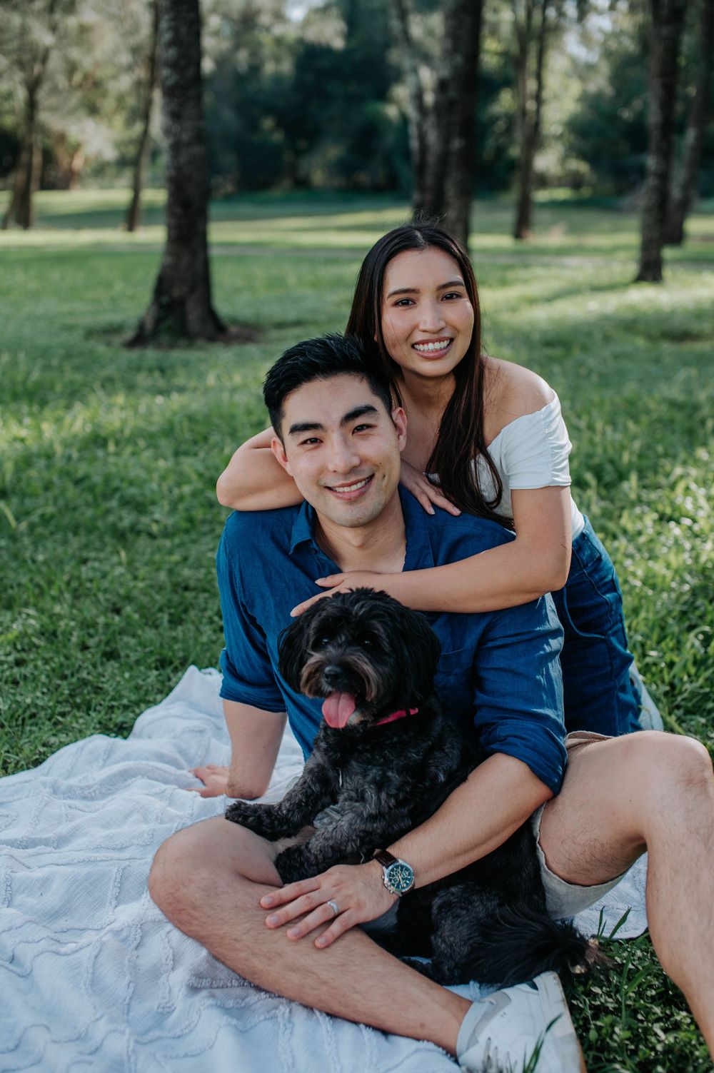 Happy couple poses with their black dog on a blanket during an outdoor photoshoot.